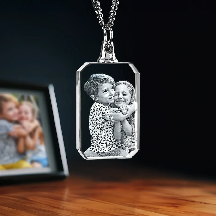 Rectangular crystal photo necklace with 2D laser-engraved image of two children hugging, hanging from a silver chain on a dark background.